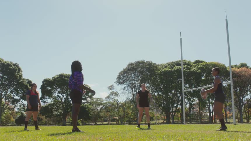 A group of female rugby players practice tossing a rugby ball on an athletics field during the day