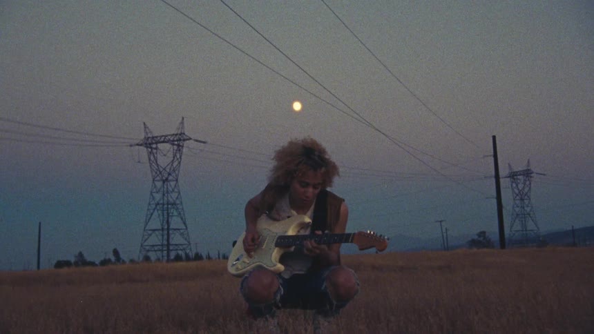 A girl is in a field playing an electric guitar and the moon is overhead and there are utility towers in the distance