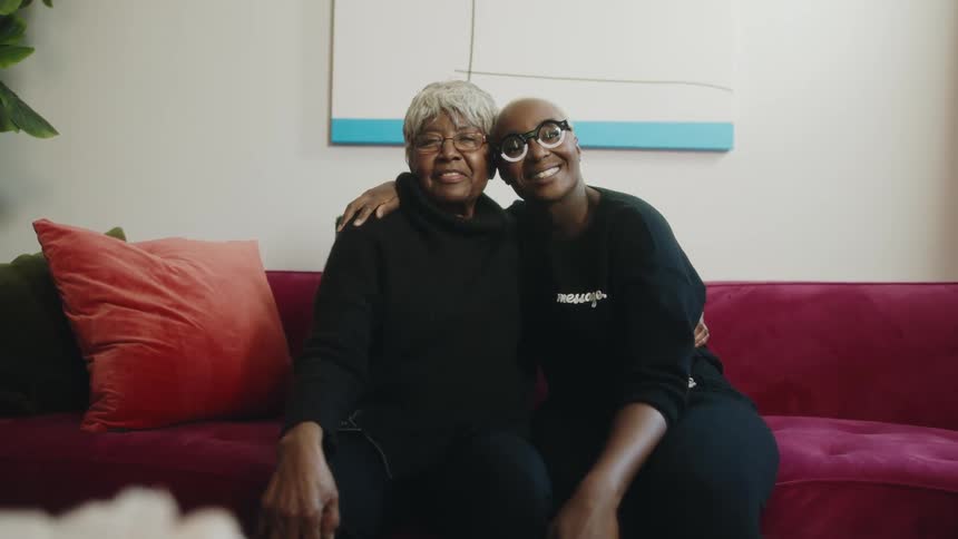 A mother and daughter smile into the camera while embracing on a sofa in the living room of a house