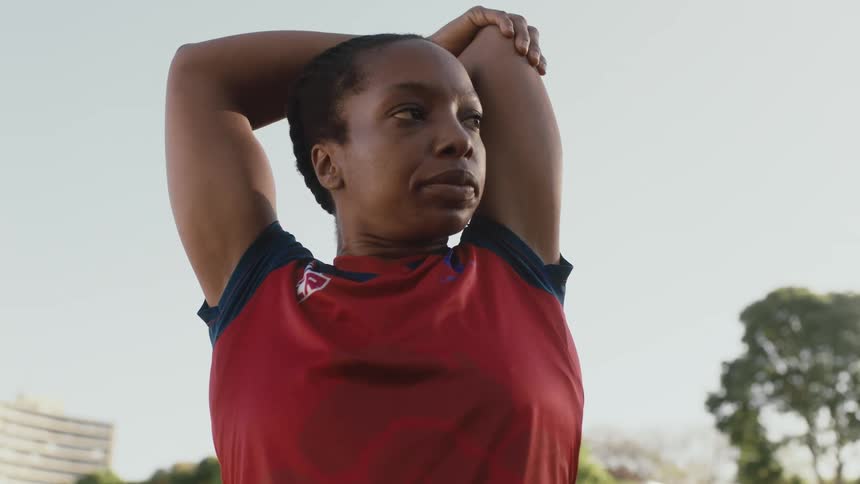 A female athlete performs stretches and exercises while standing outdoors during the day