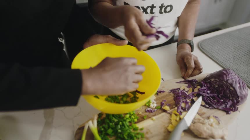 A pair of people take vegetables from a wooden cutting board and place them in a plastic bowl