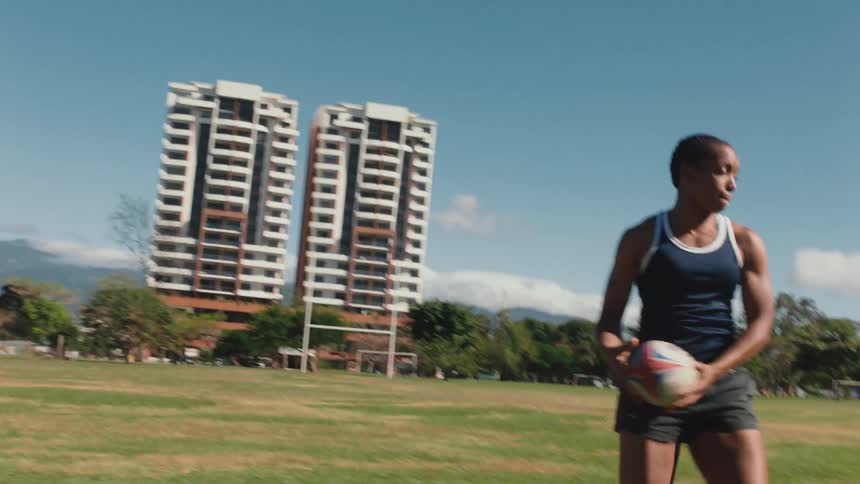 A group of women practice passing a rugby ball while running through an athletics field during the day