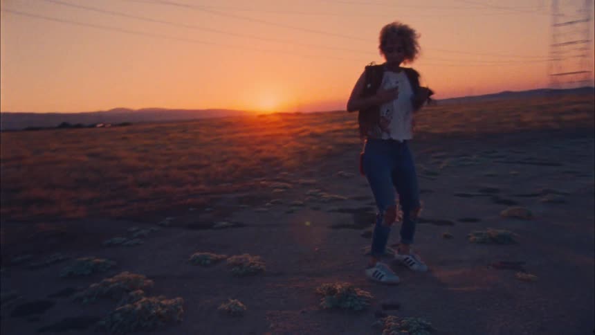 Female dancer dances on scrub land as sun sits red horizon near transmission towers and power lines