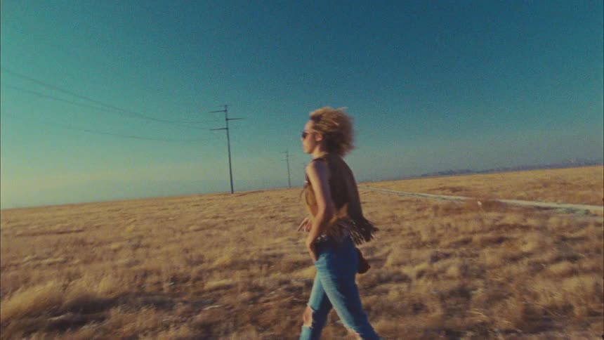 A woman exits a car parked on a dirt road through a grassland and begins to walk through the grass