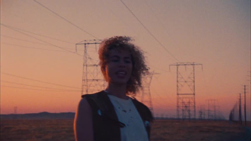 Female dancer dances on road in countryside near transmission towers standing in rows against sky tinted red by setting sun
