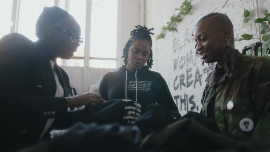 A group of women talk with one another will examining clothing together in a room during the day