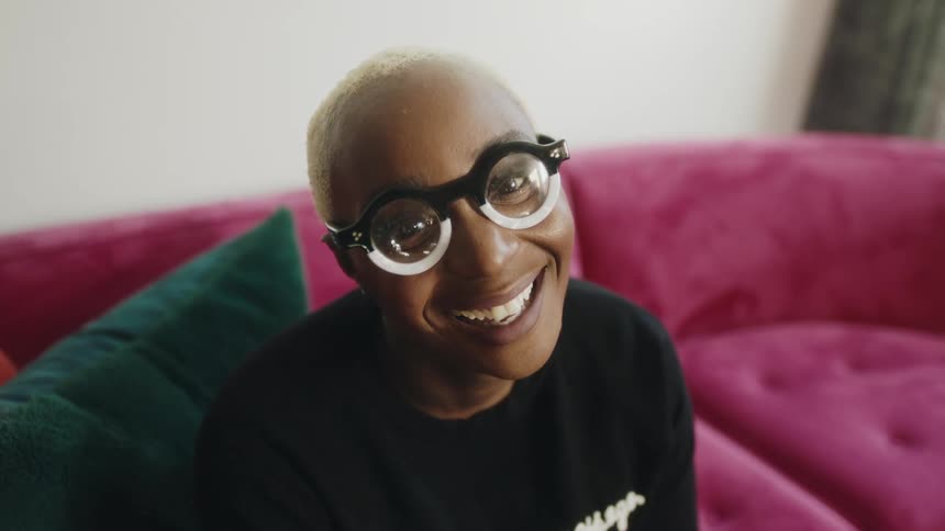 Woman with short hair thick circle glasses nods and smiles at camera sitting on pink couch