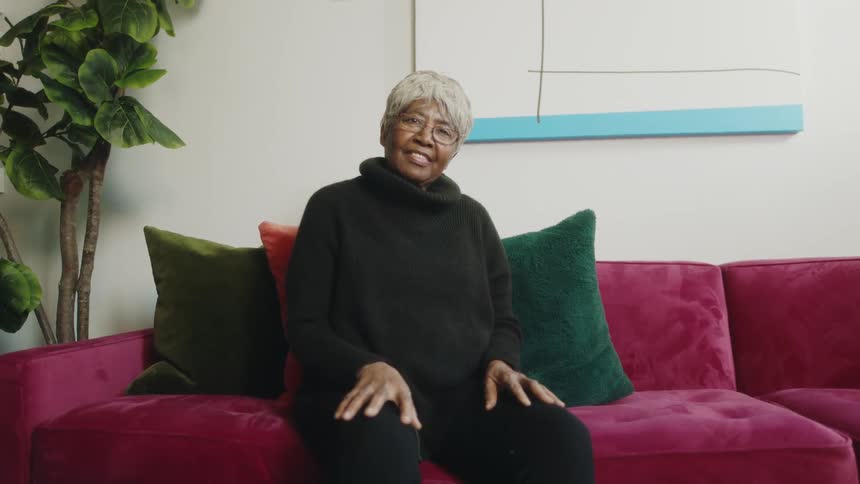 An elderly woman smiles into the camera while sitting on a sofa