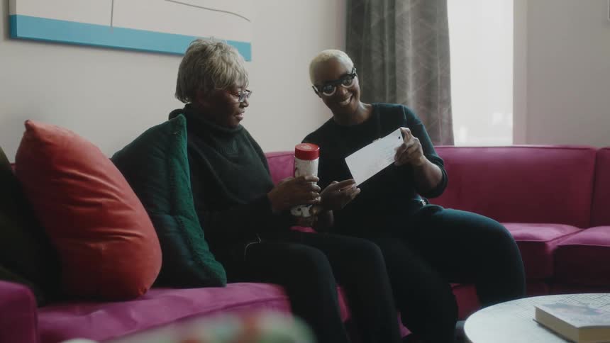 A woman reads a paper to a friend while they sit on the couch
