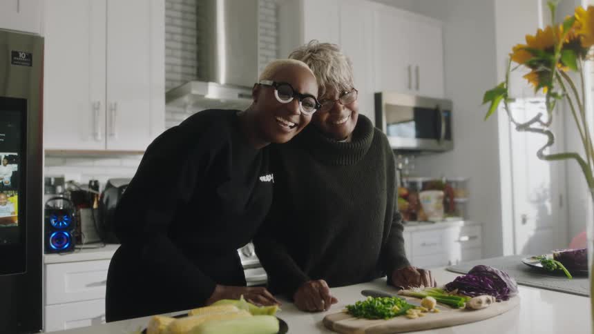 Two woman are standing and smiling in a kitchen in front of a counter top with food ingredients