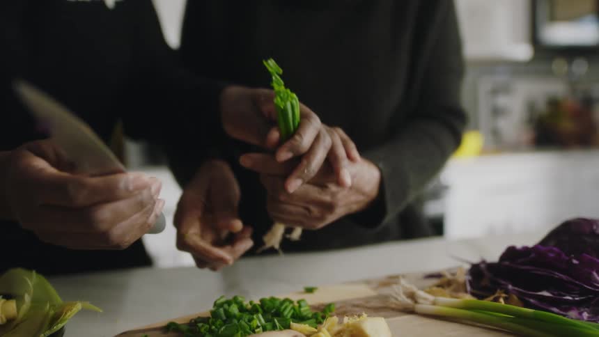 A pair of women chop vegetables together on a wooden cutting board in a home kitchen