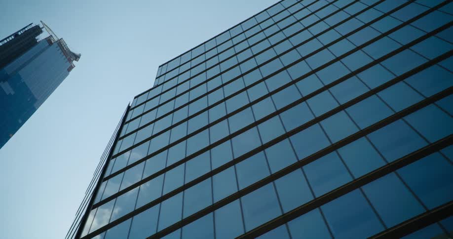 The reflections of nearby buildings and the sky shine on the surface of a tall downtown building