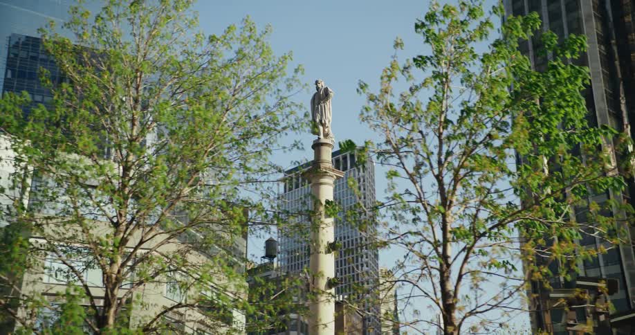A sculpture of a man sits on a tall narrow tower below tall skyscrapers