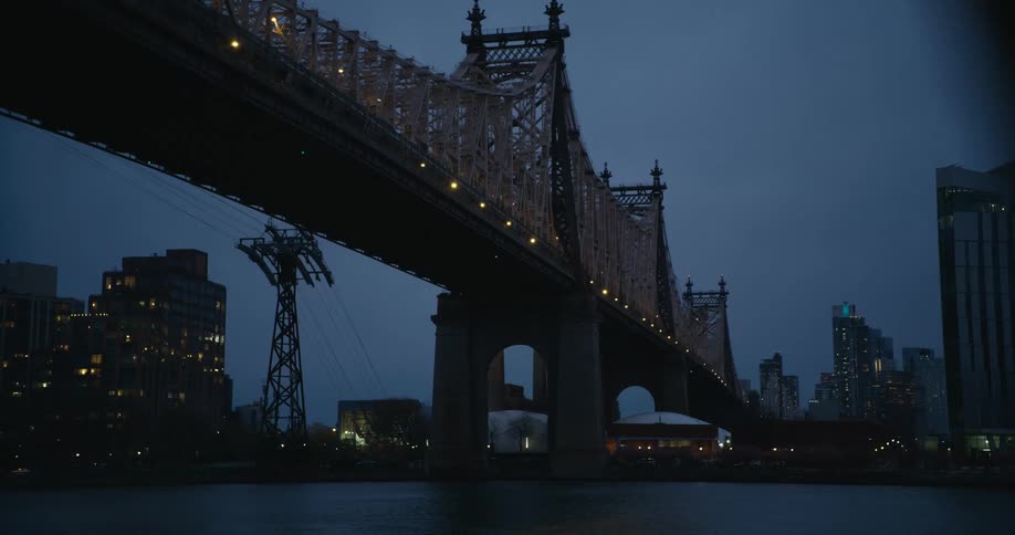 Vehicle passes by suspension bridge over river next to illuminated buildings at dusk