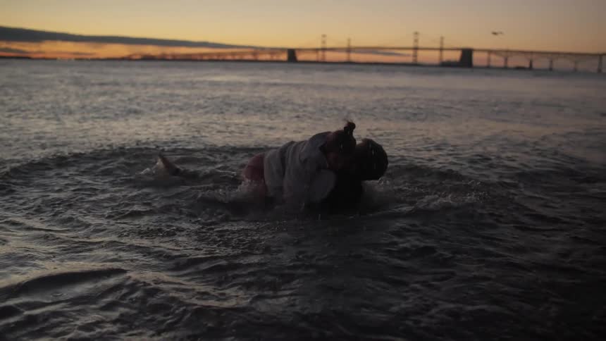 A man wades into the water carrying a woman and they laugh as they both fall back