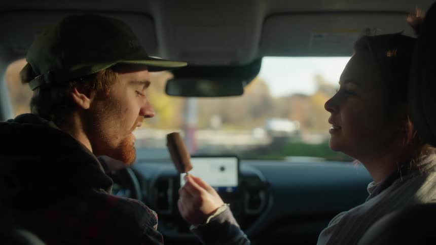 Woman shares ice cream with man with cap and beard driving car on highway