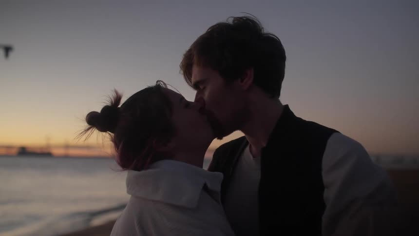 A man and woman kiss while standing on a beach at sunset