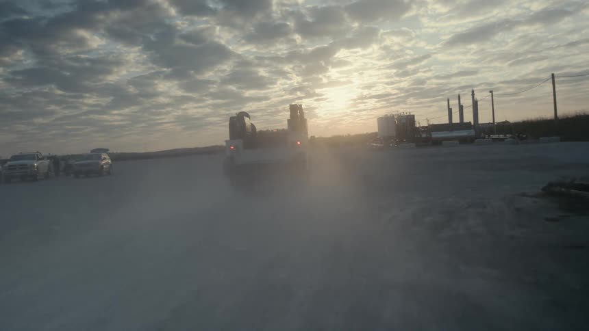 Utility truck drives over dirt road bordered by storage tanks with sun sitting near the horizzon