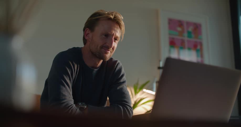 Middle aged man with beard gray sweatshirt and watch makes video call on laptop in living room
