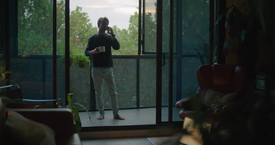 Man with sweatshirt and jeans holds cup and talks on smartphone on the balcony