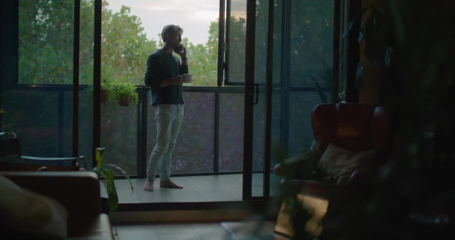 A barefoot man sips from his coffee cup while taking a phone call on the balcony of a house during the day