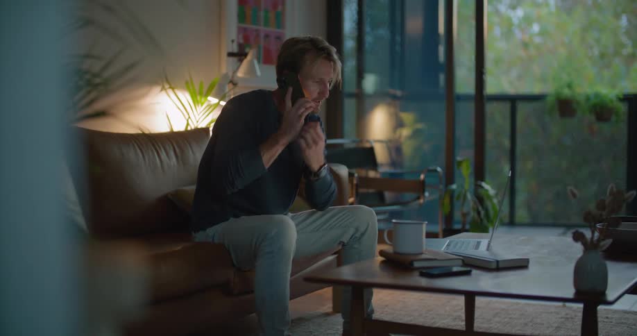 A man takes a seat on a sofa in the living room of a house and types on a laptop for a moment before holding a phone to his ear while looking through a book
