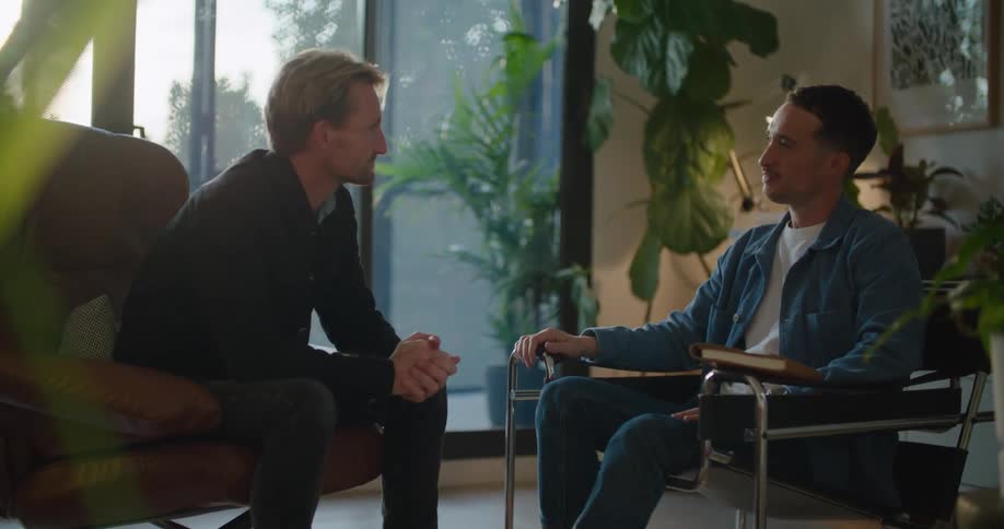 A man takes a seat across from another man and the two shake hands before beginning to chat