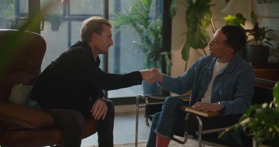 Two men are sitting in chairs talking and they shake hands and one man stands and walks away