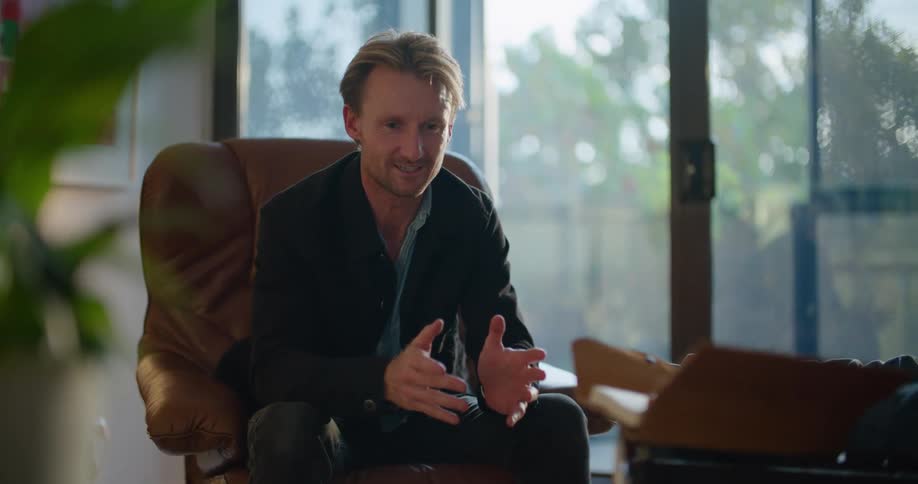 A man sits and gestures as he converses with another man seated across from him