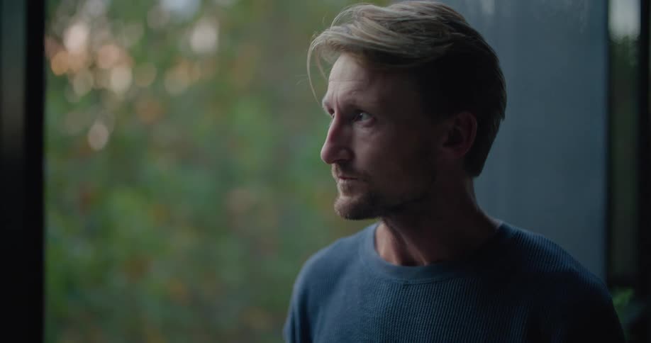 A man drinks from a cup while gazing contemplatively out of a large window during the day