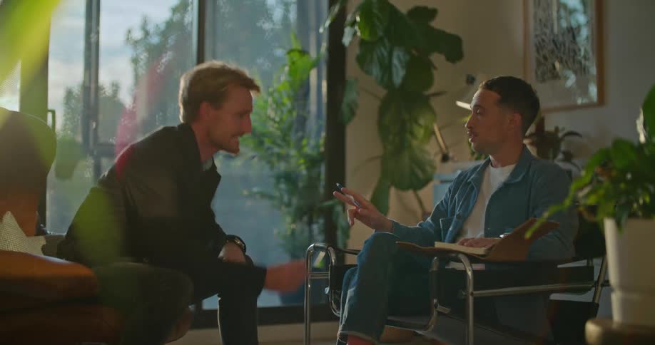 Two men are sitting in chairs talking and one man gets angry and points at the other man and knocks the book off his chair and storms out of the room