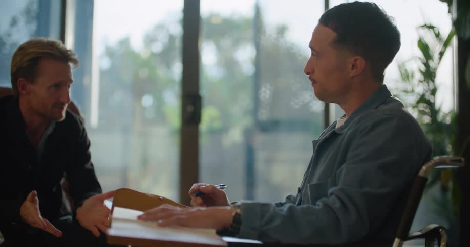 A man sitting indoors speaks to a younger man taking notes across from him