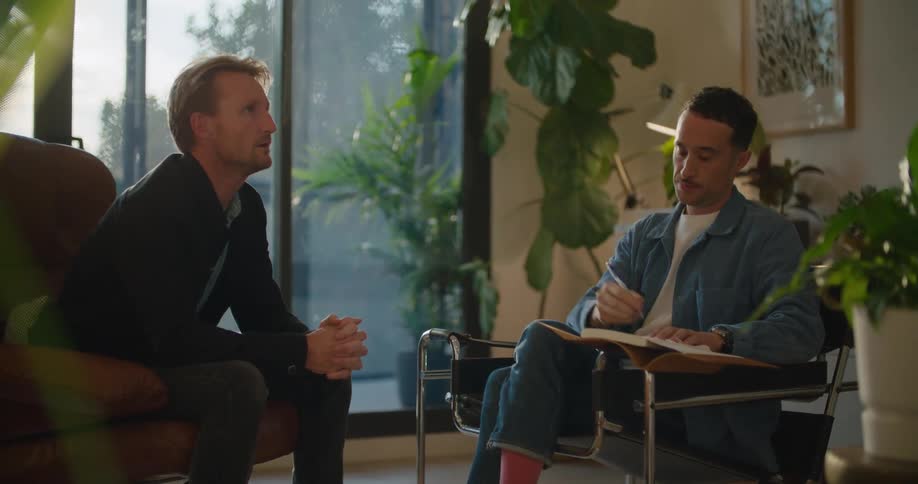 Two men are sitting in chairs talking and one man has a book open and a pen in his hand as he talks