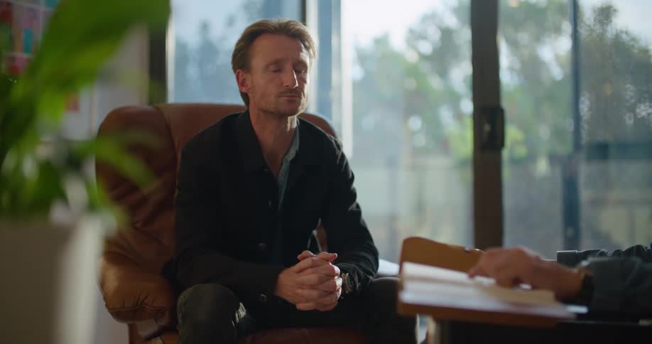 A man takes a deep breath and rubs his face with his hand while sitting and listening to a therapist