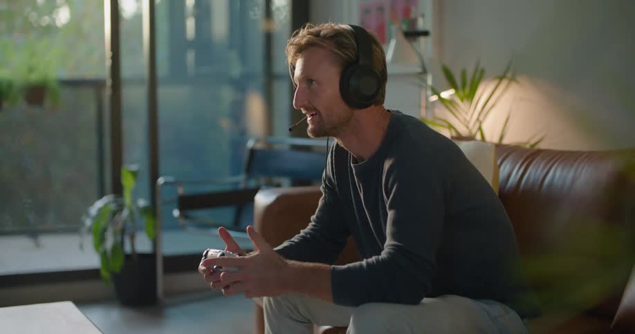 A man sitting on a leather sofa playing a video game takes off his headset and walks away