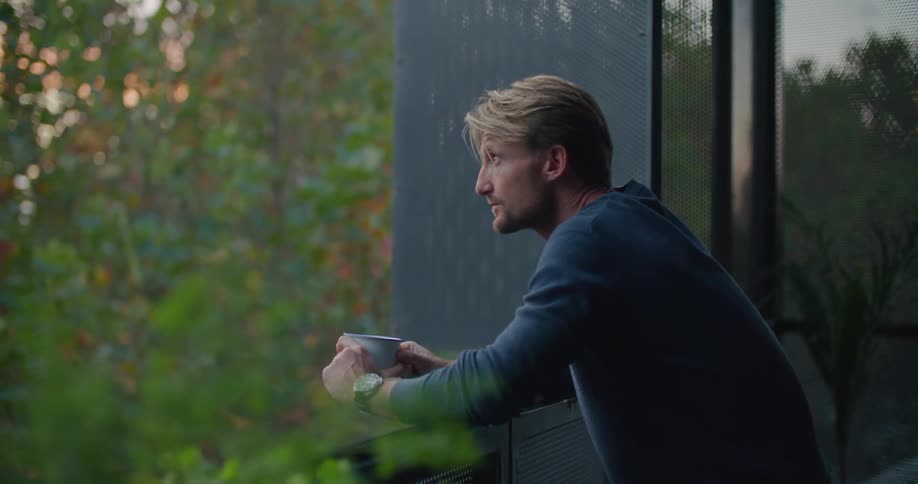 A man is standing outside on a balcony holding a coffee cup and looking around and he goes back inside