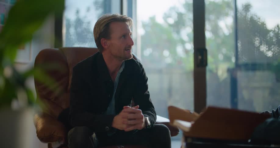 A man is sitting in a chair talking to someone and moving his hands and clasping them together