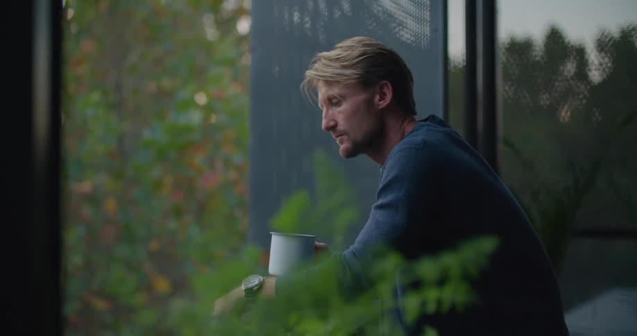 A man is standing outside on a balcony looking around and drinking from coffee cup and then he goes back inside