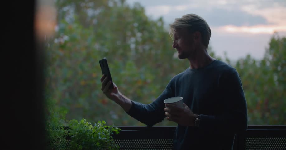 A man holds up his smartphone during a video on his balcony