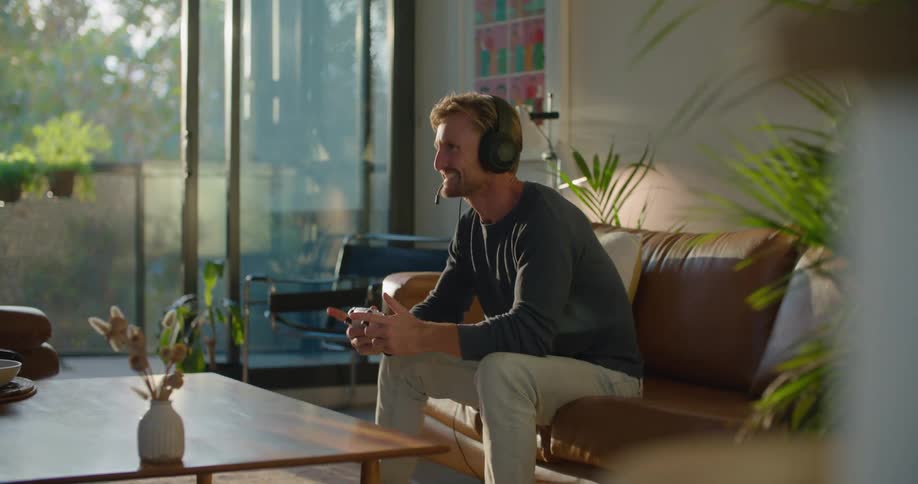 Blonde man with headphones gray sweatshirt and white pants sits on brown couch playing video game in living room at home