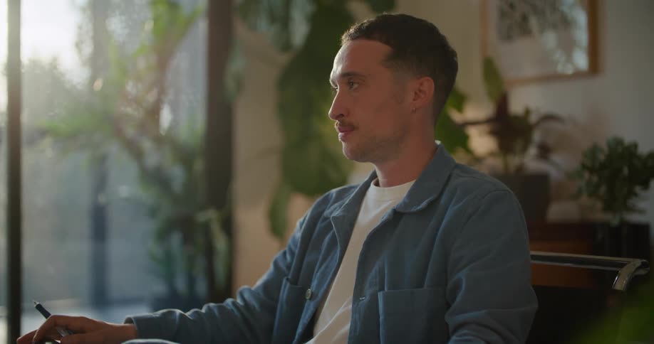 A man is sitting in a chair with a pen and a book talking to someone as he writes and holds up the book so they can see it