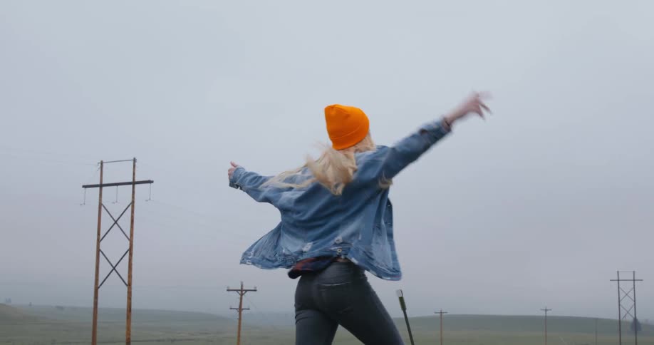A teenage girl wearing an orange knit cap and a denim jacket is dancing with her arms in the air as she moves down a street with utility poles in the distance