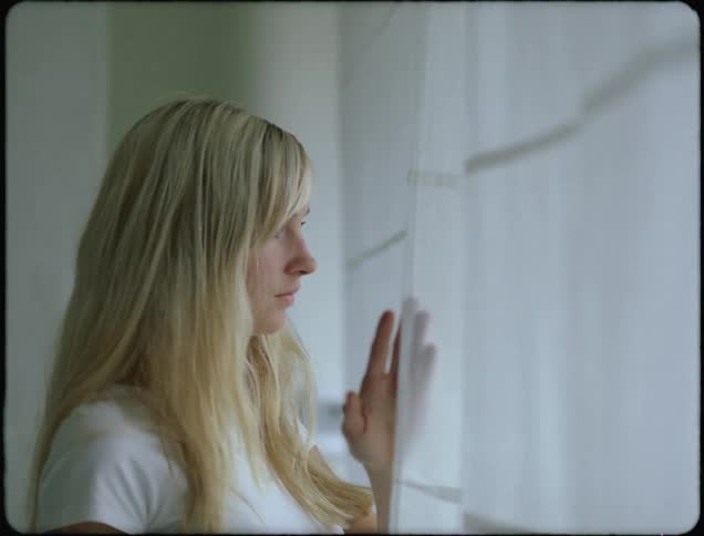 A young woman parts the curtains to gaze through a window during the day