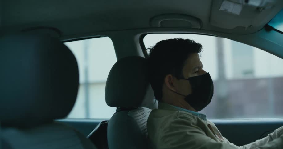 A man wearing a face mask is driving an automobile in the daytime