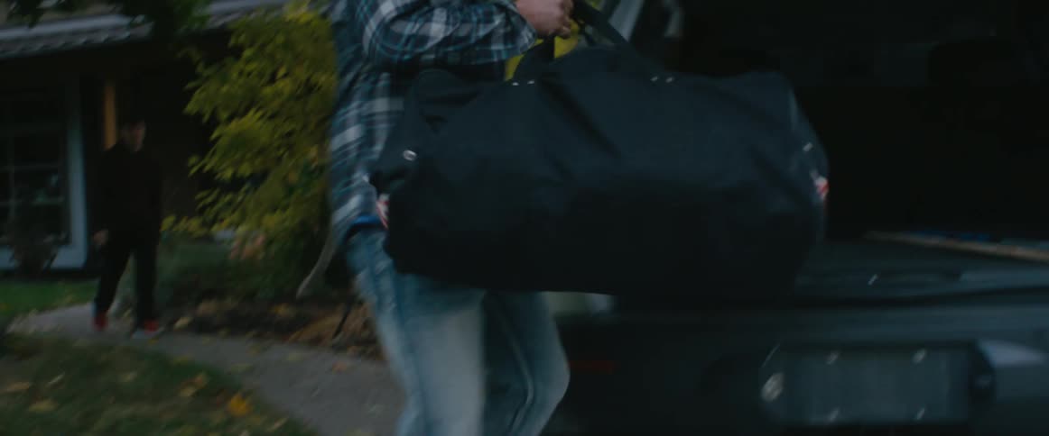 A man carries a duffel bag from a house and places it in the back of a vehicle parked in the driveway