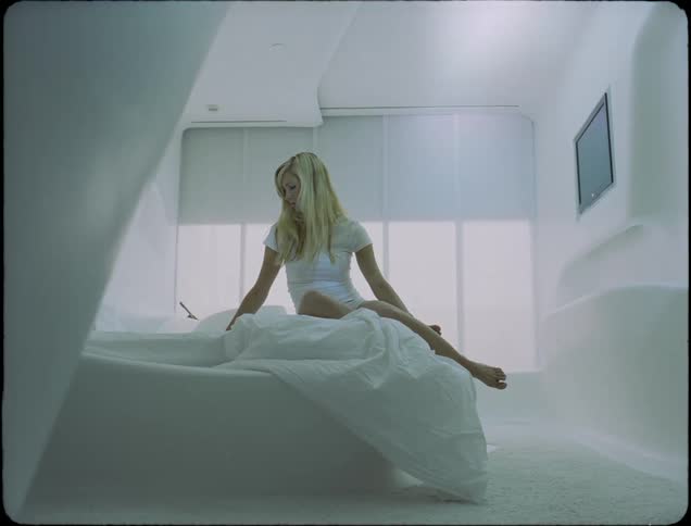 A young woman sits on a bed as automated window shades rise on the windows behind her