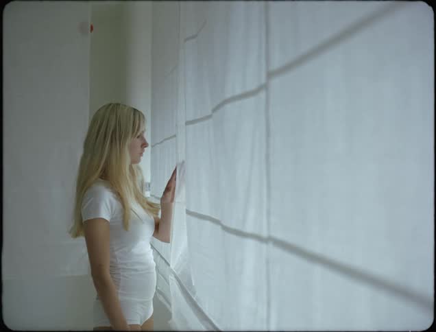 A teenage girl with long blond hair walks to windows and moves white curtains to look outside