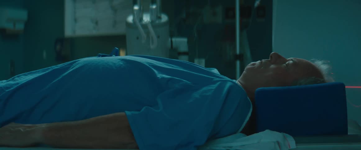 Someone is pushing a man lying on a bed and wearing a blue hospital gown in a hospital room