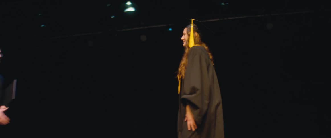 A woman gets on stage wearing a graduation cap and gown and a man hands her a black folder as they shake hands