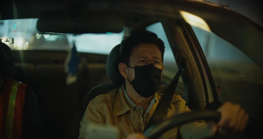 A man is wearing a face mask and driving a car and someone is sitting in the passenger seat